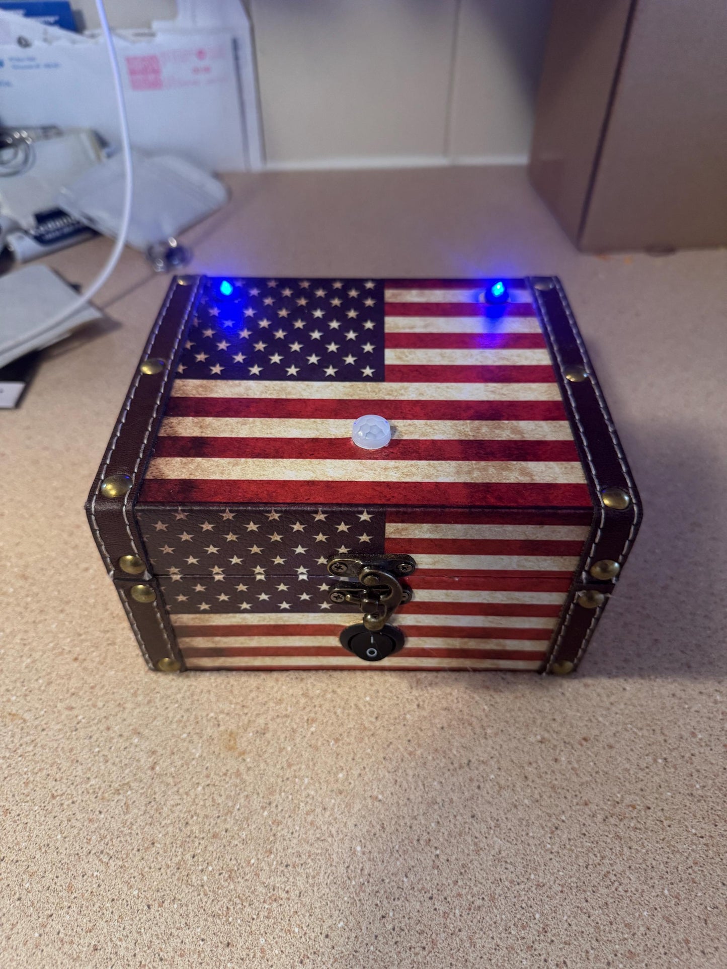 May include: A small wooden box with an American flag design. The box has a brown leather trim and metal accents. There are two blue LED lights on the top of the box and a white button in the center. There is a black switch on the bottom of the box.