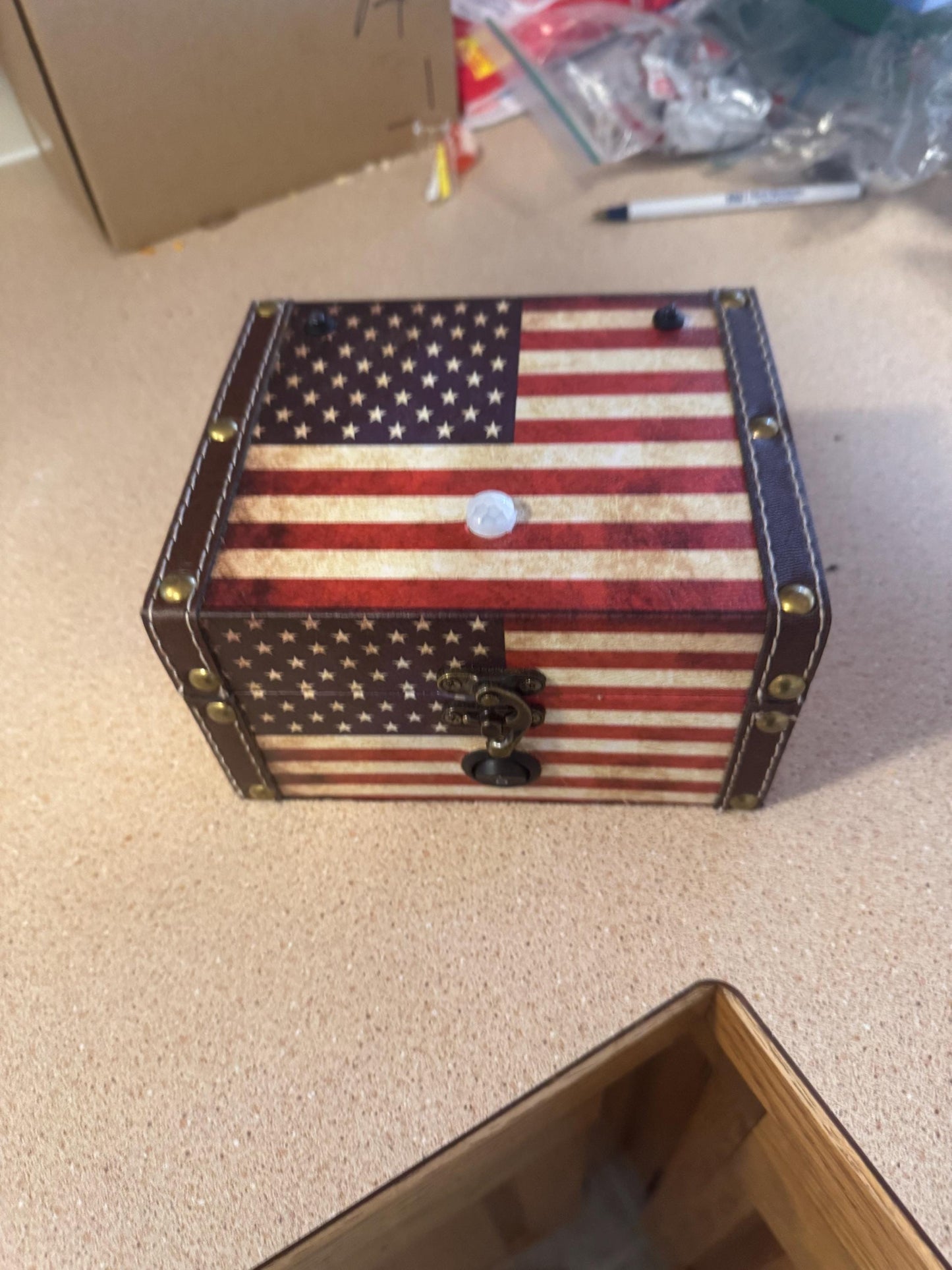 May include: A small wooden box with a brown leather exterior and a latch. The box is decorated with a vintage-style American flag design with red and white stripes and blue stars.