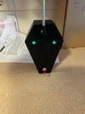 May include: A black, coffin-shaped device with two green lights and a red button. The device has a silver rod extending from the top.