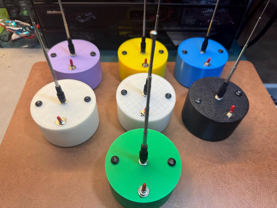 May include: Six small, round, colorful devices with antennas and red buttons. The devices are arranged in a circle on a brown surface. The devices are white, green, black, blue, yellow, and purple.