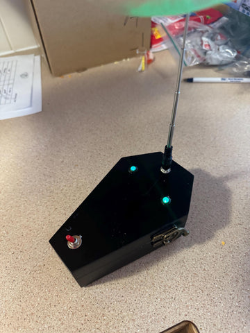 May include: A black coffin-shaped device with a silver antenna, two green lights, and a red switch. The device has a silver emblem on the side.