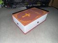 May include: A closed book shaped like a bible with a brown cover, gold lettering that reads 'HOLY BIBLE', and a gold cross on the front. The book has a white spine and is sitting on a gray surface.