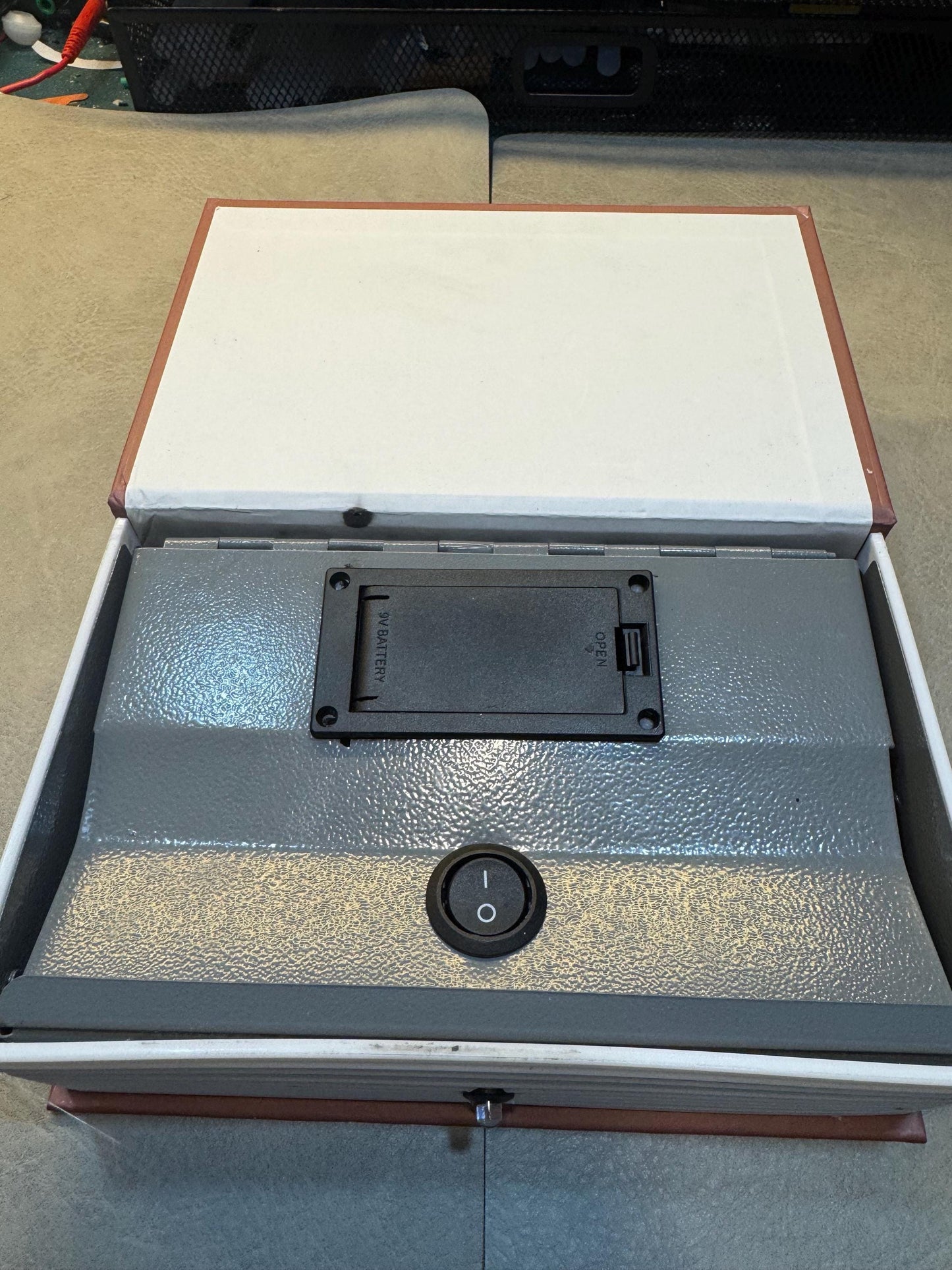 May include: A gray book safe with a black battery compartment labeled 'Battery' and a black switch labeled 'Open'. The safe has a black power switch on the front.