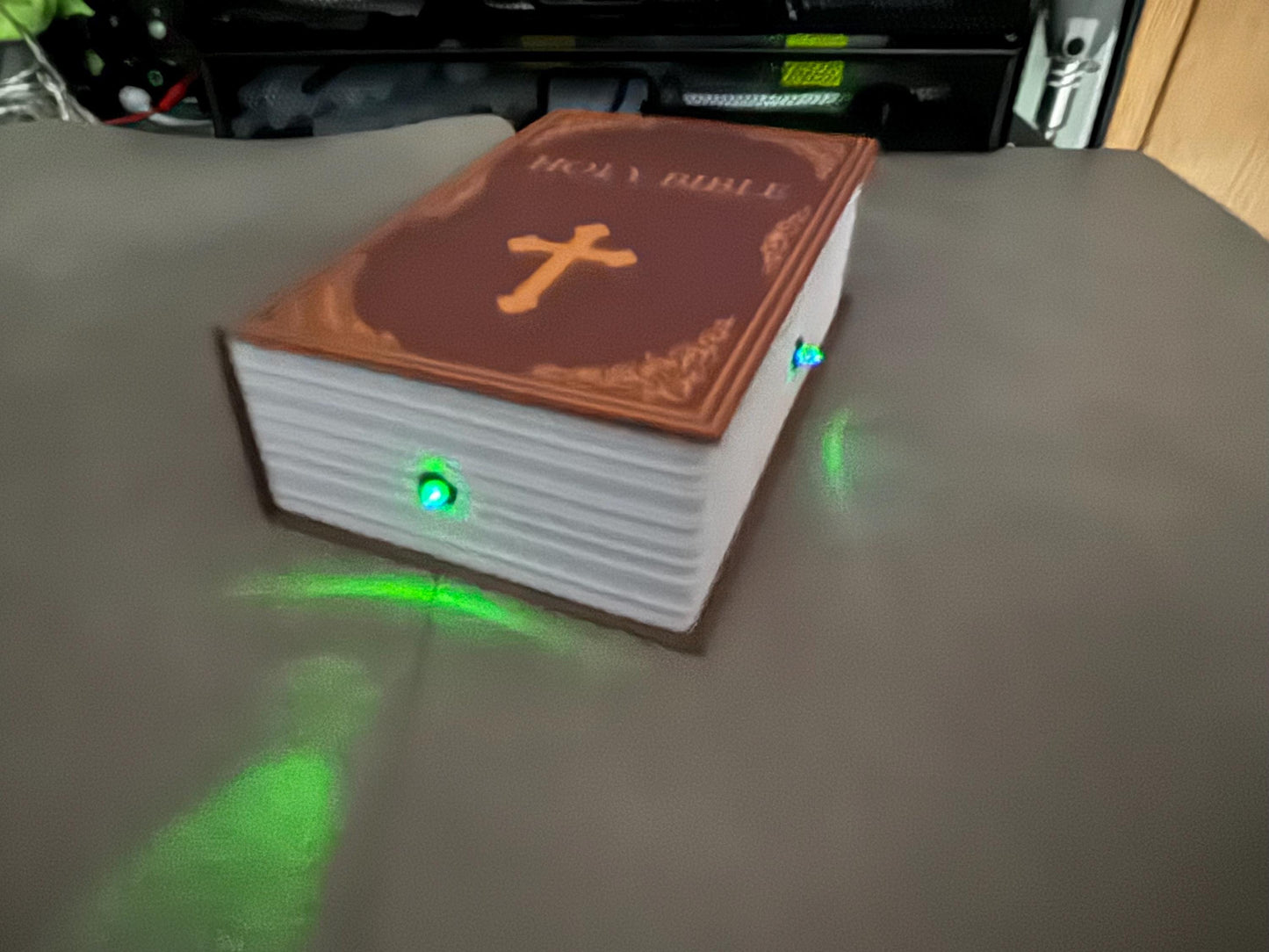 May include: A brown book with the words 'HOLY BIBLE' embossed on the cover. The book has a white cross on the front and is illuminated with green lights on the side.