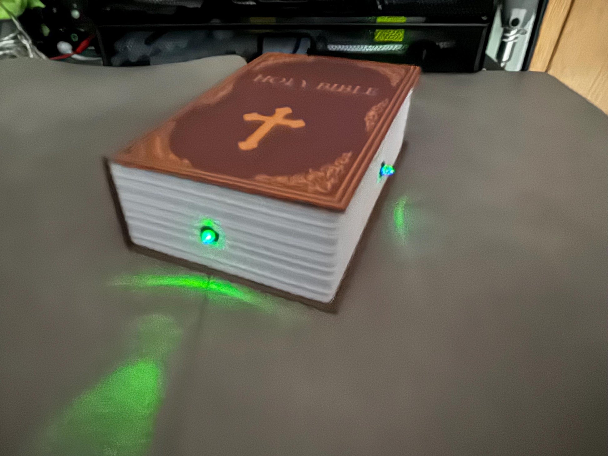 May include: A brown book with the words 'HOLY BIBLE' embossed on the cover. The book has a white cross on the front and is illuminated with green lights on the side.